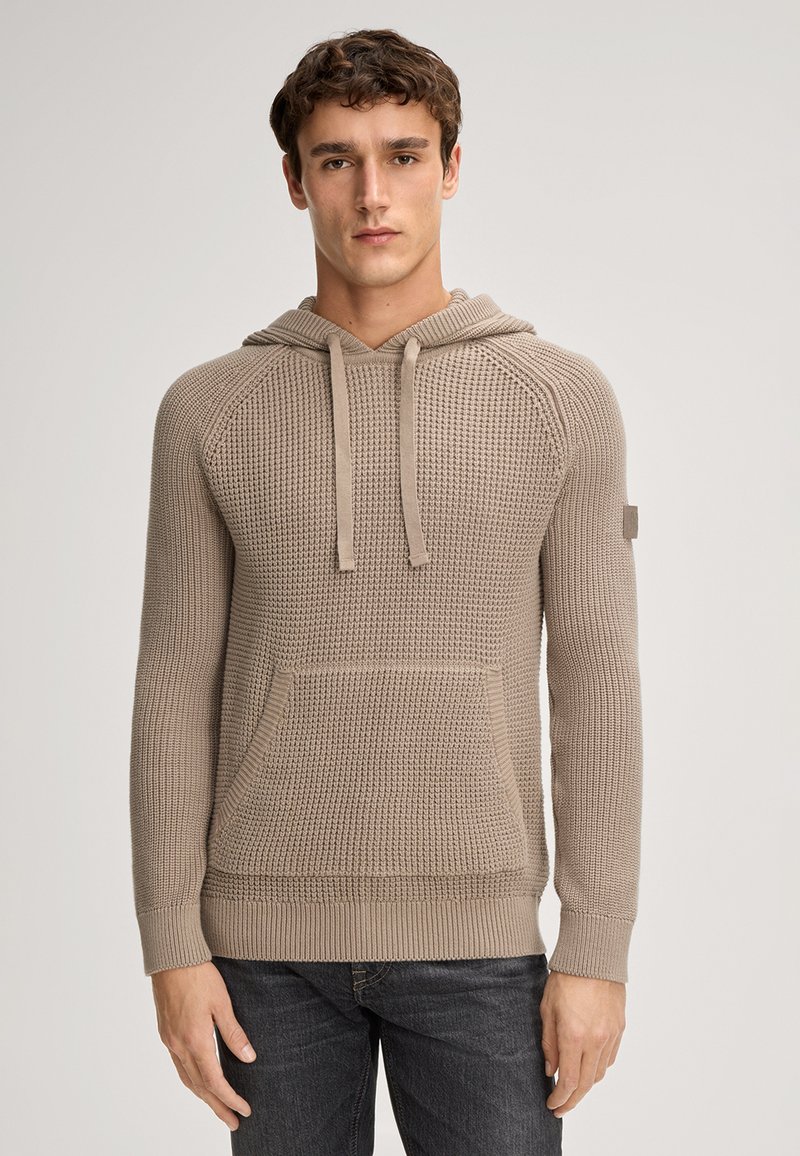 Haut à capuche tissé beige avec une finition texturée, poche kangourou, capuche avec cordons de serrage et poignets côtelés, présenté sur un modèle devant un fond neutre.