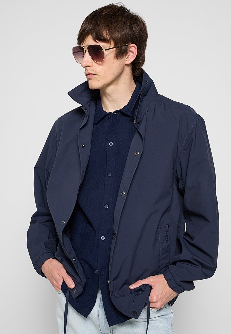 Marineblaue Windjacke mit hohem Kragen, Druckknöpfen und elastischen Bündchen über einem dunkelblauen Hemd mit Knöpfen. Kennzeichnet sich durch eine glatte, leichte Textur.