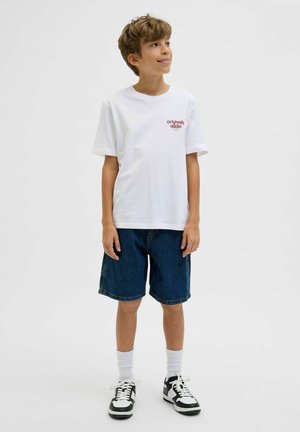 Niño sonriendo, lleva una camiseta blanca con el texto "Originals Studio", pantalones cortos vaqueros, calcetines blancos y zapatillas negras y blancas, de pie sobre un fondo blanco.