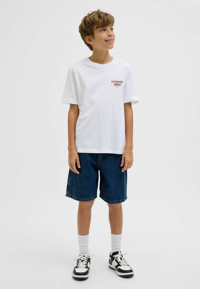 Garçon souriant, portant un t-shirt blanc avec l’inscription "Originals Studio", un short en jean, des chaussettes blanches et des baskets noires et blanches, debout sur un fond blanc.