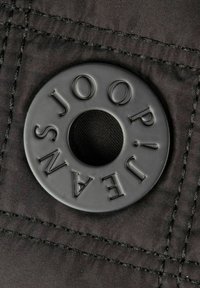 Tessuto nero con un'etichetta rotonda in plastica nera rialzata, con testo inciso "JEANS JOOP!" e foro centrale. Dettaglio delle cuciture visibile.