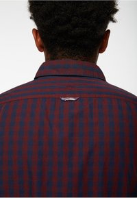 Camisa a cuadros roja y marina, con cuello abotonado y un pequeño desgarro en la parte posterior cerca del cuello, fabricada en tejido entrelazado.