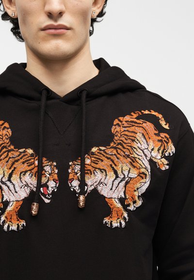 Gros plan d'une personne portant un sweat à capuche noir avec deux tigres orange brodés se faisant face sur la poitrine.
