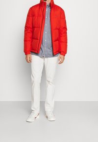 Chaqueta de plumón roja con cierre de cremallera y cuello alto, combinada con una camisa de botones azul clara y pantalones blancos. Zapatillas blancas con detalles rojos.