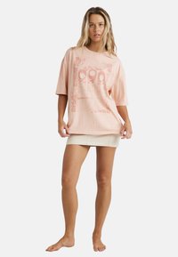 Lyserød oversized T-shirt med grafisk design og tekst, korte ærmer, blødt bomuldsstof, båret med en hvid mini-nederdel.