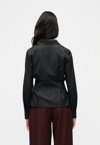 Γυναίκα που φοράει μαύρο πουκάμισο με ριγέ μοτίβο και μακριά μανίκια με γιακά, εμφανιζόμενη από πίσω, συνδυασμένο με σκούρα κόκκινα παντελόνια φαρδιάς γραμμής.