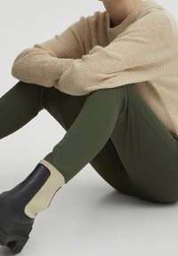 Beige gebreid trui gecombineerd met olijfgroene leggings en zwarte enkellaarzen met een beige elastische detail. Zachte texturen en aangesloten ontwerp.