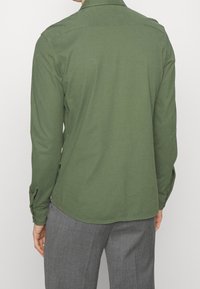 Zelená dlouhá košile vyrobená z lehkého materiálu, s špičatým límcem a knoflíkovými manžetami, spárovaná s šedými kalhotami s jemnou texturou.