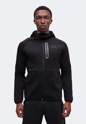 Homme portant un sweat à capuche noir zippé avec une poche poitrine, debout devant un fond clair uni.