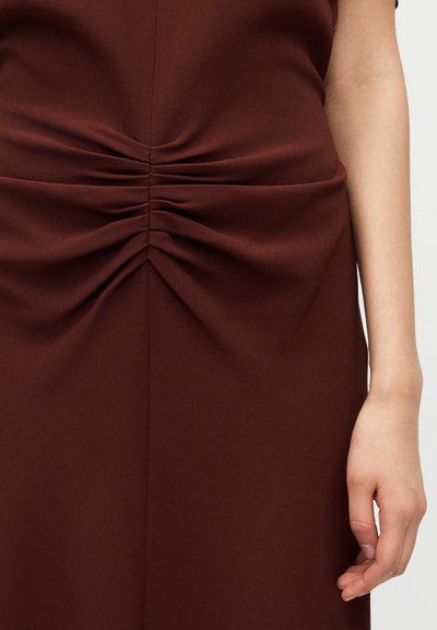 Μπέργκουντι, εφαρμο στό φόρεμα με πιέτες μπροστά και υφή τελειώματος. Κοντινή λήψη υφάσματος με ορατό χέρι στο πλάι.