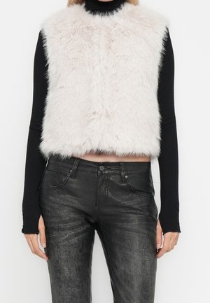 Personne portant un gilet sans manches en fausse fourrure blanche sur un haut noir à manches longues avec un pantalon en cuir noir texturé.