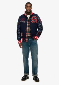 Chaqueta varsity de la marina con acentos rojos, logos bordados y puños a rayas, llevada sobre una camisa de cuadros y combinada con vaqueros azules y zapatos negros.