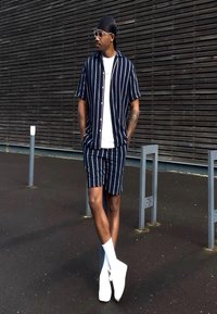 Άνδρας με μαύρο durag, γυαλιά ηλίου, μπλε ναυτικό ριγέ πουκάμισο και σορτς, λευκό φανελάκι, κάλτσες και παπούτσια, στηρίζεται σε μια κολόνα έξω.