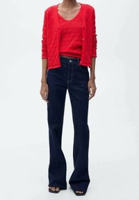 Rød strikket cardigan over en rød tekstureret top, parret med flagrende mørk denim jeans. Sætningen har en tætsiddende stil og flade sandaler.