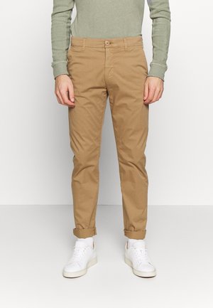 Mężczyzna ubrany w beżowe spodnie chinos podwinięte u dołu, jasnozieloną koszulę z długim rękawem oraz białe trampki, stojący na jednolitym białym tle.