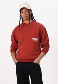 Rød genser med ribbestrikkede mansjetter og hem, med en frontlomme og hvit tekstlogo. Modellen har på seg en beige caps og svarte bukser.