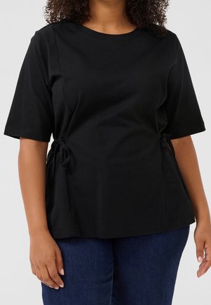 Nainen, jolla on musta lyhythihainen paita sivusolmujen kanssa ja tummansiniset farkut, seisoo yksinkertaisen vaalean taustan edessä.