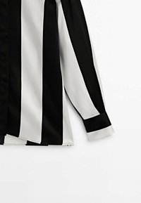Chemise à manches longues avec larges rayures verticales noires et blanches, montrant la moitié inférieure et la manche droite sur fond blanc.