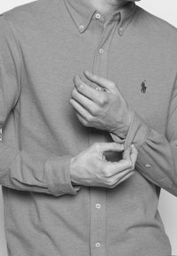 Camicia grigia con colletto, caratterizzata da un piccolo logo ricamato. Il tessuto appare morbido con una trama testurizzata. Le maniche vengono regolate.