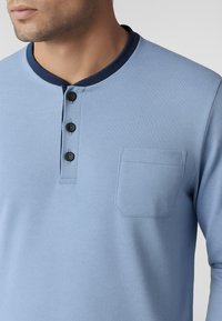 Camicia a maniche lunghe azzurra chiara con colletto blu navy, tre bottoni neri e una tasca sul petto di forma rettangolare. Materiale in cotone.