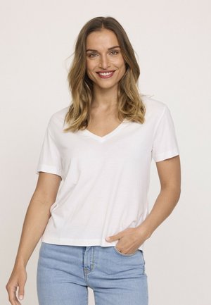 T-shirt en coton blanc avec un col en V et des manches courtes, offrant une coupe décontractée. Porté avec un jean bleu clair, affichant un style décontracté.