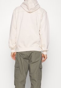 Sudadera con capucha de color beige claro y corte relajado, con puños acanalados y un bolsillo tipo canguro. Combinada con pantalones cargo de color verde oliva que cuentan con bolsillos laterales.