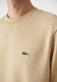 Sudadera de algodón beige con cuello redondo y un pequeño logo bordado de cocodrilo verde en el pecho izquierdo. Textura suave.