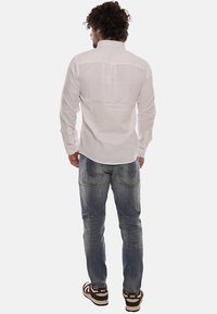 Camicia bianca a maniche lunghe con colletto, abbinata a jeans blu sbiaditi e sneakers marroni, vista da dietro. Texture liscia, design casual.