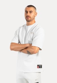 Witte t-shirt met korte mouwen en een ronde hals, voorzien van een subtiel logo en een patch met tekst en accenten aan de linkerkant. Zachte stoftextuur.