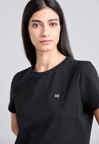 Juodas medvilnės marškinėlis su apvalia iškirpte ir trumpomis rankovėmis, ant kairės krūtinės pusės yra auksinė logotipo etiketė.
