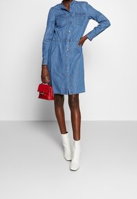 Farkkupaitamekko, jossa pitkät hihat, napitettava etuosa, kaksi rintataskua ja joustava vyötärö. Yhdistetty punaisen käsilaukun ja valkoisten nilkkureiden kanssa.