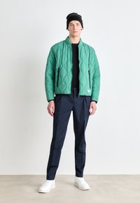 Veste bomber matelassée verte avec col montant, chemise noire à manches longues, pantalon bleu marine et baskets blanches. Le mannequin porte un bonnet noir.