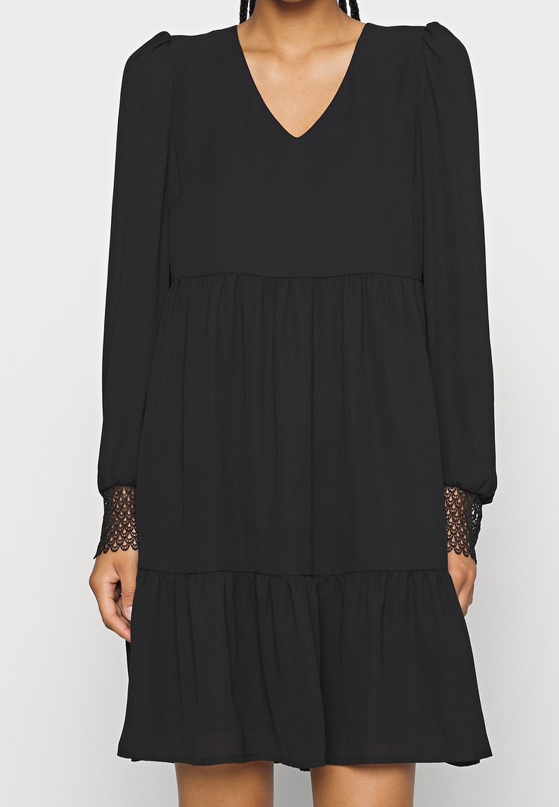 Femme portant une robe noire à manches longues avec col en V, jupe à étages, épaules bouffantes et poignets en dentelle, debout devant un fond uni.