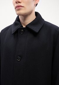 Cappotto nero in tessuto texturizzato, con colletto a revers, quattro grandi bottoni e vestibilità sartoriale. Accentuato da una sottile intrecciatura diagonale.