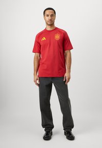 Κόκκινο αθλητικό μπλουζάκι με κοντά μανίκια, με κίτρινες λεπτομέρειες και έμβλημα της Ισπανίας. Φοριέται με σκούρα τζιν και μαύρα παπούτσια.