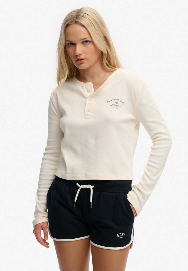 Ung kvinde iført en cremefarvet langærmet henley-trøje og sorte atletikshorts med bindebånd og hvide kanter, stående med den ene hånd i lommen.