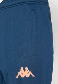 Φόρμες σε ναυτικό μπλε χρώμα από λείο ύφασμα, με ροζ λογότυπο που απεικονίζει δύο καθιστές φιγούρες. Διαθέτει πλαϊνές τσέπες και ελαστική μέση.