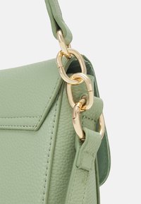 Gros plan sur une sangle de sac à main en cuir texturé vert clair, avec de grandes maillons de chaîne dorée brillante reliant la sangle au sac.