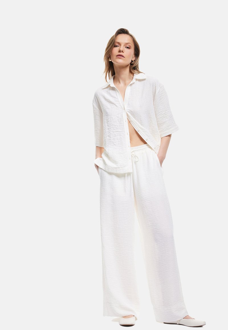 Camisa blanca de textura, con mangas cortas, cierre de botones y pantalones sueltos con cordón a juego. Diseño simple sin patrones ni acentos visibles.