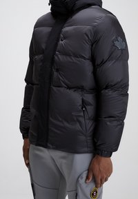 Doudoune noire avec un col haut, des boutons-pression et un emblème en relief en forme de feuille d'érable sur l'épaule gauche. Matière lisse et brillante.