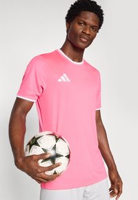 Ροζ αθλητικό μπλουζάκι με λευκή τρέσα, με μικρό λευκό λογότυπο Adidas. Το μοντέλο κρατάει μία μαύρη και λευκή μπάλα ποδοσφαίρου με σχέδιο.