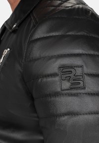 Zwarte leren jas met doorgestikte schouderpanelen, een ritszak en een geprint logo-patch op de linkermouw. Soepele textuur.