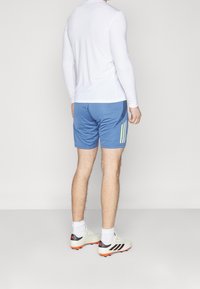 Urheiluasu, joka koostuu istuvasta valkoisesta pitkähihanaisesta paidasta ja sinisistä shortseista, joissa on keltaiset raidat, yhdistettynä valkoisiin ja mustiin jalkapallokengän rautoihin.