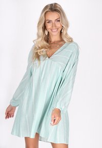 Rochie de un teal deschis cu mâneci lungi și puffate, decolteu în V și dungi diagonale albe. Material moale, cu o croială lejeră și talie strânsă.