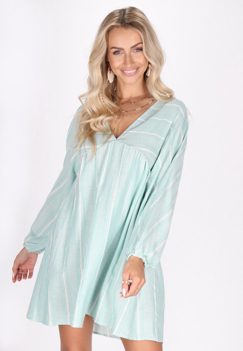 Rochie de un teal deschis cu mâneci lungi și puffate, decolteu în V și dungi diagonale albe. Material moale, cu o croială lejeră și talie strânsă.