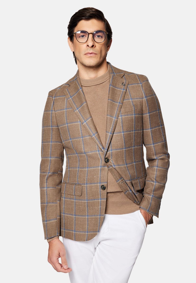 Brązowa marynarka w kratę z niebieskimi akcentami, o dopasowanym kroju i klapach typu notch. Nosi się ją na beżowym swetrze o fakturze, w zestawieniu z białymi spodniami.