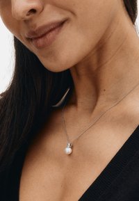 Sølvkæde med rundt perlevedhæng med en dekorativ sølvkappe, hvilende på hud synlig i en sort beklædning.