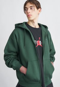 Sweatshirt verde com fecho éclair, feito de tecido suave, apresentando um capuz com cordão, dois bolsos frontais e punhos canelados. Usado sobre uma tshirt preta.