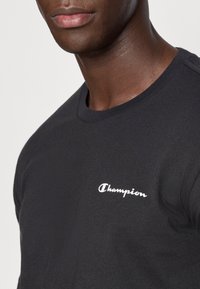 Κοντινό πλάνο ενός ατόμου που φοράει μαύρο μπλουζάκι με στρογγυλή λαιμόκοψη και μικρό λευκό λογότυπο "Champion" στο αριστερό στήθος.