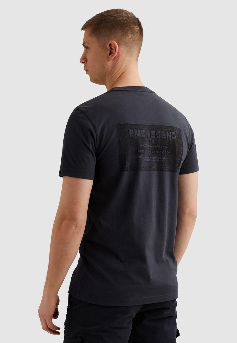 Homme aux cheveux courts portant un t-shirt PME Legend bleu marine foncé avec un logo imprimé dans le dos, debout devant un fond clair uni.